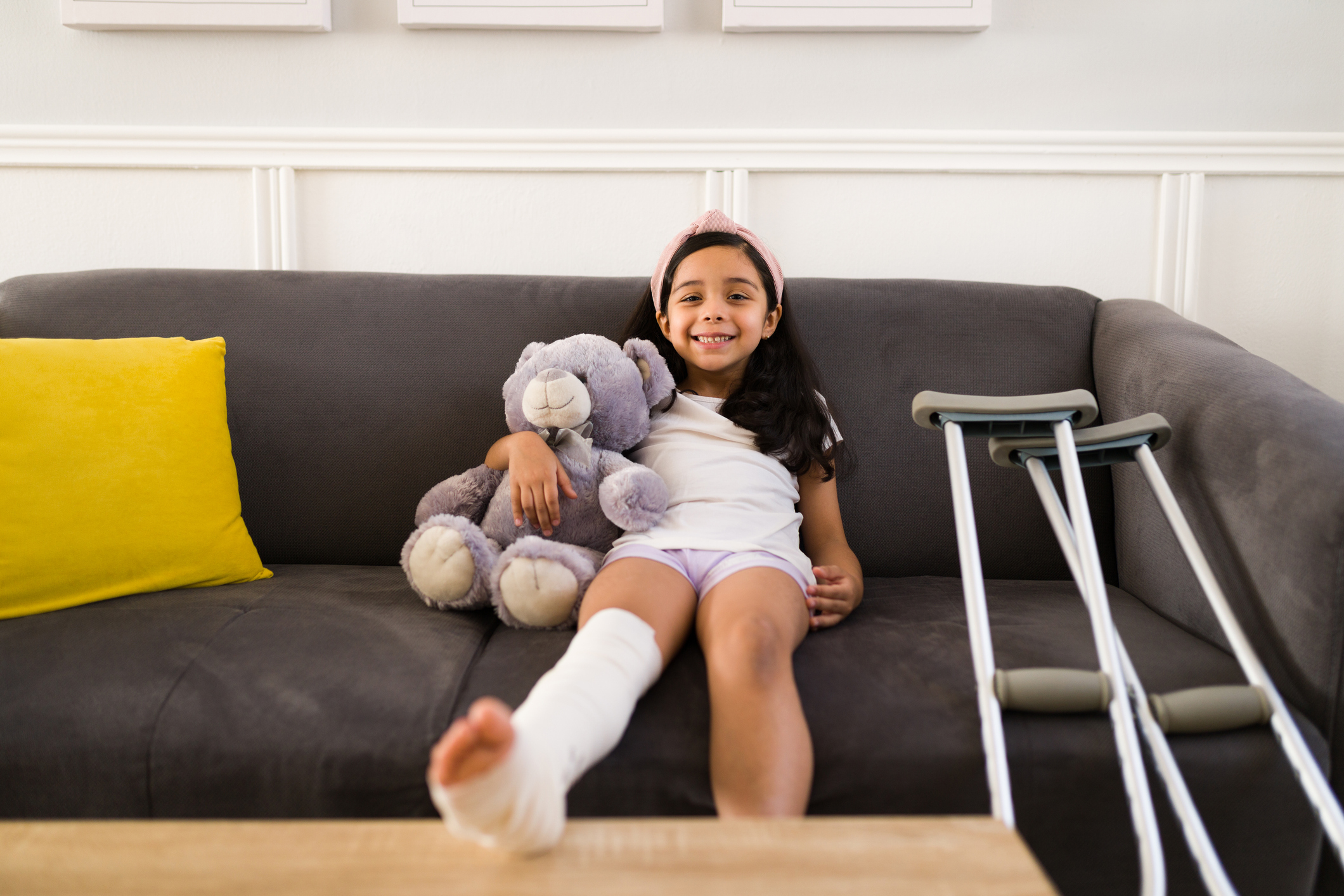 School-aged girl on couch holding stuffy, crutches at side. Wearing leg cast.