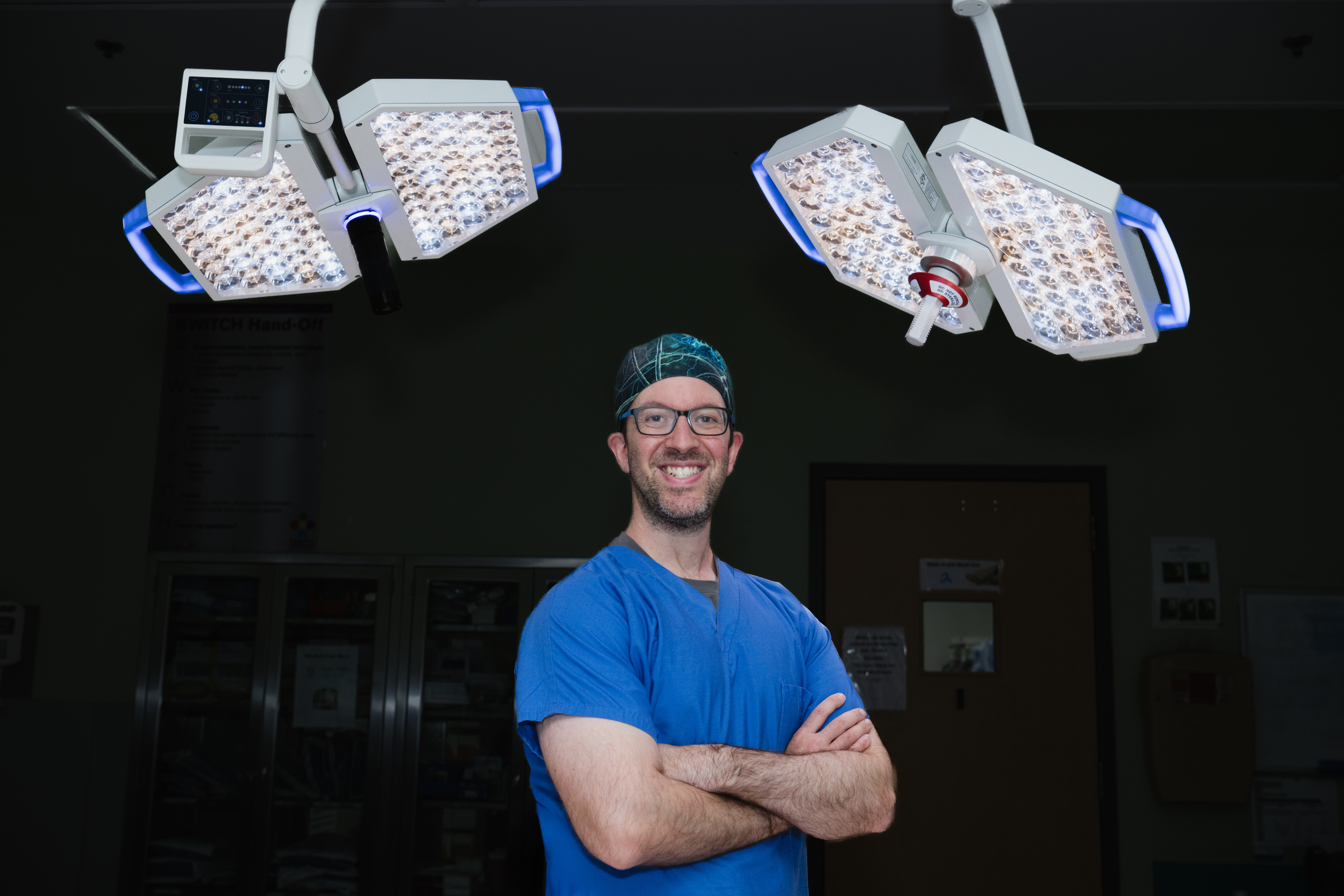 Dr. Hersh, neurosurgeon, stands in the operation room with is arms crossed, with operating room lights behind him. Dr. Hersh, neurosurgeon, stands in the operation room with is arms crossed, with operating room lights behind him.