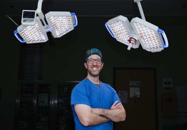 Dr. Hersh, neurosurgeon, stands in the operation room with is arms crossed, with operating room lights behind him. Dr. Hersh, neurosurgeon, stands in the operation room with is arms crossed, with operating room lights behind him.