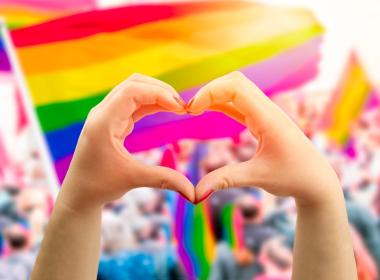 Supporting hands make heart sign and wave in front of a rainbow flag flying on the sidelines of a summer Pride parade