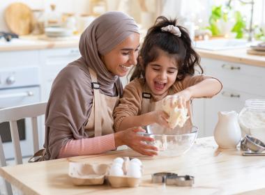 Mom and young daughter crack eggs into a bowl in kitchen, happily.