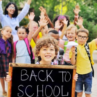 School age boy holds "Back to School" sign while friends and parents wave in the background